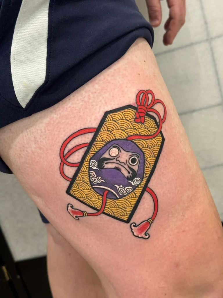 Colorful tattoo of omamori lucky charm with Daruma doll, done in Tokyo by Mangesh Rane – traditional Japanese symbols of luck and protection | TATTOOINTOKYO